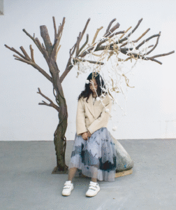 I sat under a knotted rope “tree” installation beside a stone with handprints, evoking Beckett’s Waiting for Godot and the existential act of waiting. CN: 艺术家安静坐在结绳“树”装置下，旁边是带手印的石头，呼应贝克特《等待戈多》，呈现等待的存在主义动作。